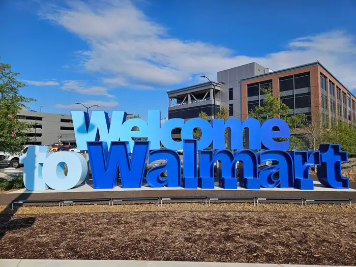 Large blue 'Welcome to Walmart' sign in a parking lot with modern buildings behind. (Informative)